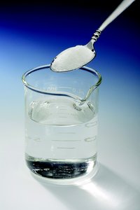 Water is the liquid in the beaker, and the white solid in the spoon is sugar.
